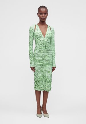 Mujer con vestido midi verde con estampado de leopardo, manga larga fruncida, con aberturas en los hombros y tacones altos beige, de pie frente a un fondo liso.