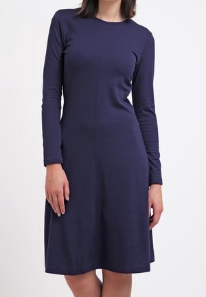 Kobieta ubrana w granatową sukienkę z długim rękawem do kolan, stojąca z rozluźnionymi ramionami na jednolitym białym tle.