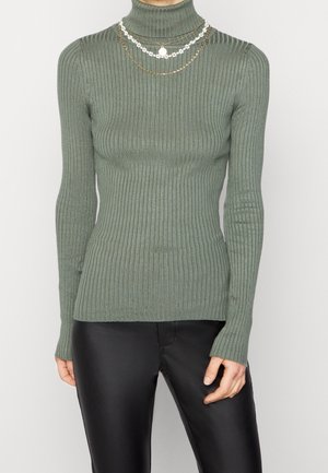 Femme portant un pull à col roulé côtelé vert associé à un pantalon en cuir noir et à des colliers floraux dorés et blancs superposés.