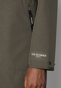 Chaqueta verde oscura con una textura suave, que cuenta con un bolsillo con cremallera, detalles de costura y el logo "ILSE JACOBSEN HORNbæk" en blanco.