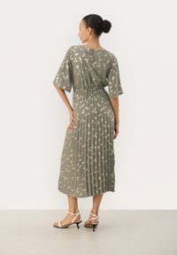 Pleatattu midi-mekko oliivinvihreänä, kevyestä kankaasta, lyhyillä hihoilla, valkoisilla abstrakteilla kuvioilla ja takana avonaista yksityiskohtaa.