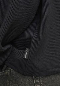 Tissu noir côtelé avec un design ajusté, comportant une étiquette visible portant la mention "ORIGINALS™" près de l'ourlet, associé à un jean gris foncé.