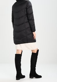 Zwarte pufferjas met chevronsteek, tot de knie, gedragen over een crèmekleurige rok en gecombineerd met zwarte suède laarzen tot de knie.