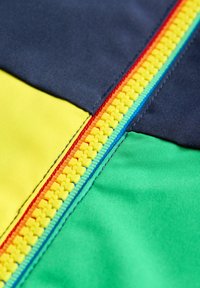 Värikäs kangas, jossa on keltaisia, laivastonsinisiä, vihreitä ja oransseja alueita, ja johon kuuluu keskeinen monivärinen vetoketju, joka korostaa muotoilua.