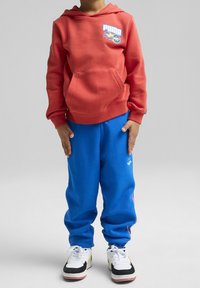 Kind trägt einen roten Puma-Hoodie mit Flammenlogo, blaue Jogginghose und weiße Turnschuhe, steht mit den Händen an den Seiten.