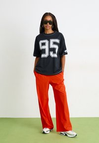 Black oversized t-shirt with white "95" graphic, paired with bright orange wide-leg pants and white sneakers with black accents.
