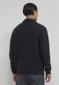 Uomo con i capelli corti scuri che indossa un maglione a coste scuro e pantaloni grigi, in piedi rivolto di spalle contro uno sfondo chiaro e neutro.