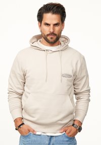 Beige cotton hoodie with drawstrings and front pocket. Features logo text on the left chest. Styled with light blue denim and a watch.