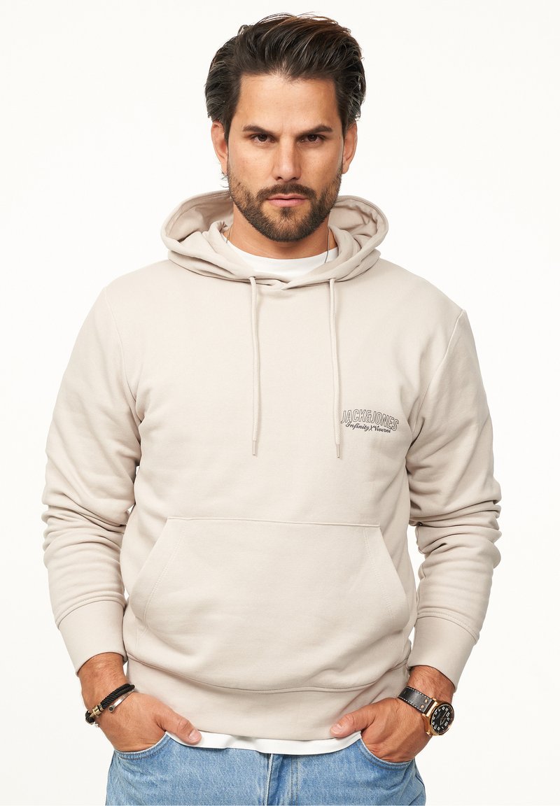 Beige cotton hoodie with drawstrings and front pocket. Features logo text on the left chest. Styled with light blue denim and a watch.