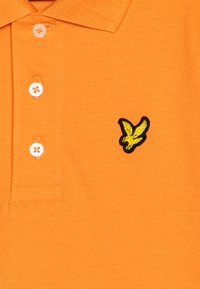 Orange polo skjorte laget av teksturert stoff, med en tre-knaps placket og en brodert gul fuglelogo på brystet.