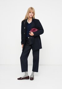 Ναυτικό σακάκι διπλής όψης, μπορντό τσάντα, σκούρες τζιν παντελόνες, γκρι κάλτσες και γυαλιστερά μπορντώ μοκασίνια. Γυναίκα με μακριά ξανθά μαλλιά.