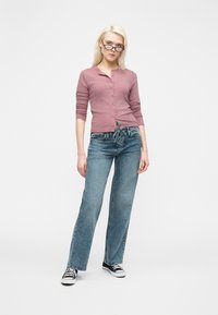 Ξανθιά γυναίκα με μαύρα γυαλιά, μωβ πουκάμισο με κουμπιά, τζιν με κορδόνια και μαύρα αθλητικά παπούτσια που στέκεται μπροστά σε απλό λευκό φόντο.