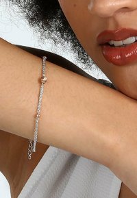 Bracciale a catena in argento con una bead in oro; collegamenti delicati e chiusura regolabile; indossato su un braccio abbronzato su uno sfondo bianco.