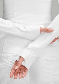 Haut blanc à manches longues avec un design ajusté, comportant des ouvertures pour les pouces dans les poignets et une texture lisse, associé à un vêtement blanc en dessous.