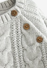 Suéter de punto en gris claro con patrones de cable texturizados y botones de madera. La tela tiene una textura suave y gruesa con detalles intrincados.