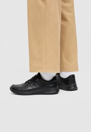 Zapatillas de cuero negras usadas con calcetines blancos y pantalones beige, mostradas de lado y por detrás sobre un fondo gris liso.