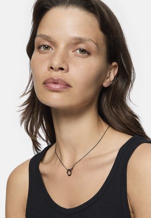 Zwarte ketting met een ronde hanger, gedragen over een zwart tanktop. De hanger heeft een gladde oppervlakte en een minimalistisch ontwerp.