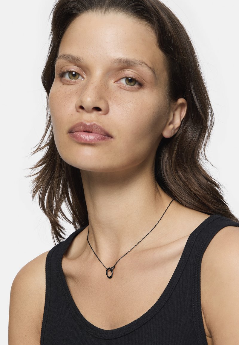 Schwarze Halskette mit einem runden Anhänger, über einem schwarzen Trägershirt getragen. Der Anhänger hat eine glatte Oberfläche und ein minimalistisches Design.
