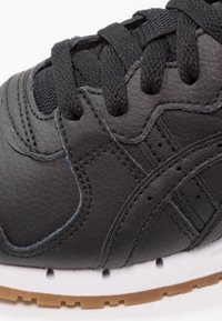 Black leather sneaker with textured surface, round toe, black laces, and white midsole, featuring visible stitching and gum rubber outsole.