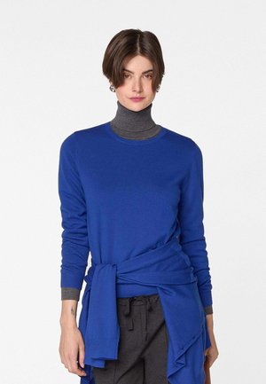 Junge Frau mit kurzen braunen Haaren, die einen blauen Pullover über einem grauen Rollkragen trägt, mit einem blauen Kleidungsstück um die Taille gebunden.