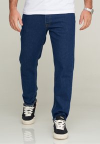 Jeans de mezclilla azul oscuro de corte recto, con costuras sutiles, bolsillos frontales clásicos, y combinados con zapatillas en blanco y negro.