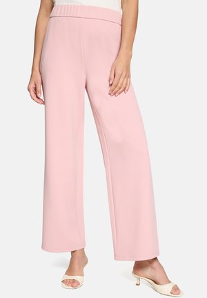 Nő magas derekú, széles szárú pasztell rózsaszín nadrágban és fehér, nyitott orrú magassarkú szandálban, sima háttér előtt állva.