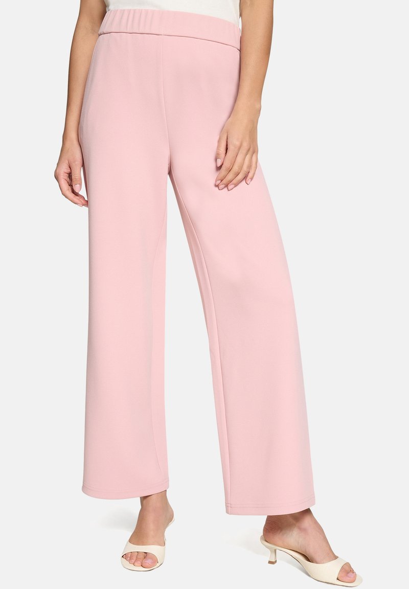 Vrouw die een pastelroze broek met hoge taille en wijde pijpen draagt, gecombineerd met witte open schoenen met hakken, staand voor een effen achtergrond.