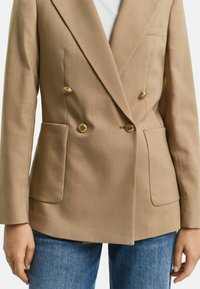 Blazer beige doppiopetto con bottoni dorati, tasche laterali e rever a punta, abbinato a jeans in denim blu. Vestibilità su misura e tessuto strutturato.