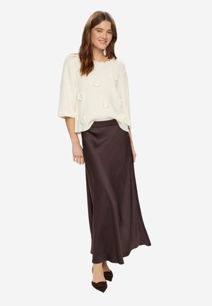 Femme portant un pull en maille crème avec des détails floraux, une longue jupe en satin marron foncé et des chaussures pointues foncées, debout sur un fond blanc.