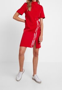 Röd klänning med korta ärmar, hög krage och slits på sidan. Har tre vita ränder längs vänster sida. Bärs med vita sneakers.