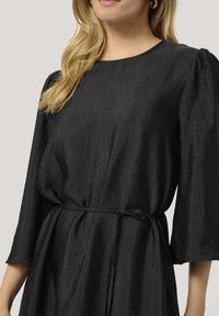 Robe noire plissée avec un col rond, des manches trois-quarts et un lien à la taille. Texture lisse avec un léger éclat.