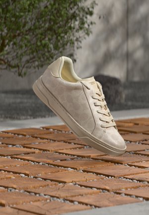 Baskets en cuir beige clair avec lacets flottant au-dessus d’une surface extérieure en bois et pierre à motifs avec de la verdure en arrière-plan.