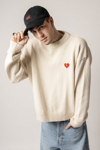 Beige knit sweater with ribbed hem and cuffs, featuring a red broken heart design on the chest. Worn with blue denim jeans and a black cap.