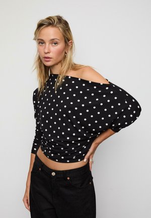 Schwarzes Off-Shoulder-Oberteil mit weißen Pünktchen, langärmlig und locker geschnitten, kombiniert mit hoch geschnittenen schwarzen Jeans.