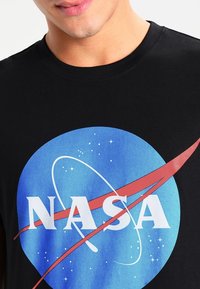 Svart T-shirt med en stor blå cirkulär NASA-logga, röd svepning, vit text och stjärnmönster på den blå bakgrunden.