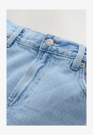 Gros plan sur un jean en denim bleu clair montrant la ceinture, le bouton en métal, les passants de ceinture et les coutures de la poche avant sur un fond uni.