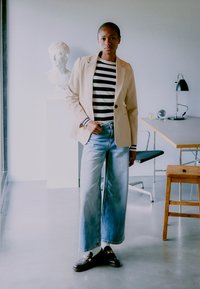 Blazer beige sobre una camiseta de rayas en blanco y negro, combinado con unos jeans de pierna ancha de color azul claro y zapatos negros, en un interior moderno.