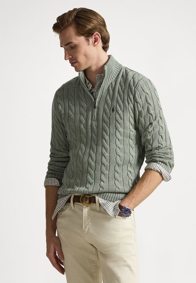 Maglione verde a trecce con collo a mezza zip, indossato sopra una camicia a quadri. Abbinato a pantaloni kaki e un orologio da polso.