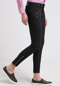 Černé slim-fit kalhoty s bočními zipy, vyrobené z pružného materiálu. Vyznačují se vysokým pasem a hladkým povrchem. V kombinaci s nazouvacími botami.