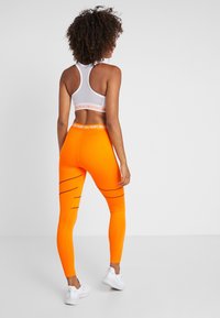 Pomarańczowe legginsy sportowe z czarnymi paskami, w połączeniu z białym stanikiem sportowym z teksturowanym elastycznym paskiem. Białe sneakersy dopełniają stylizację.