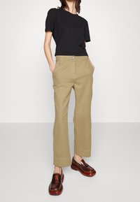 Black short-sleeve t-shirt paired with light khaki straight-leg trousers featuring front pockets and a button detail. Brown slip-on shoes.