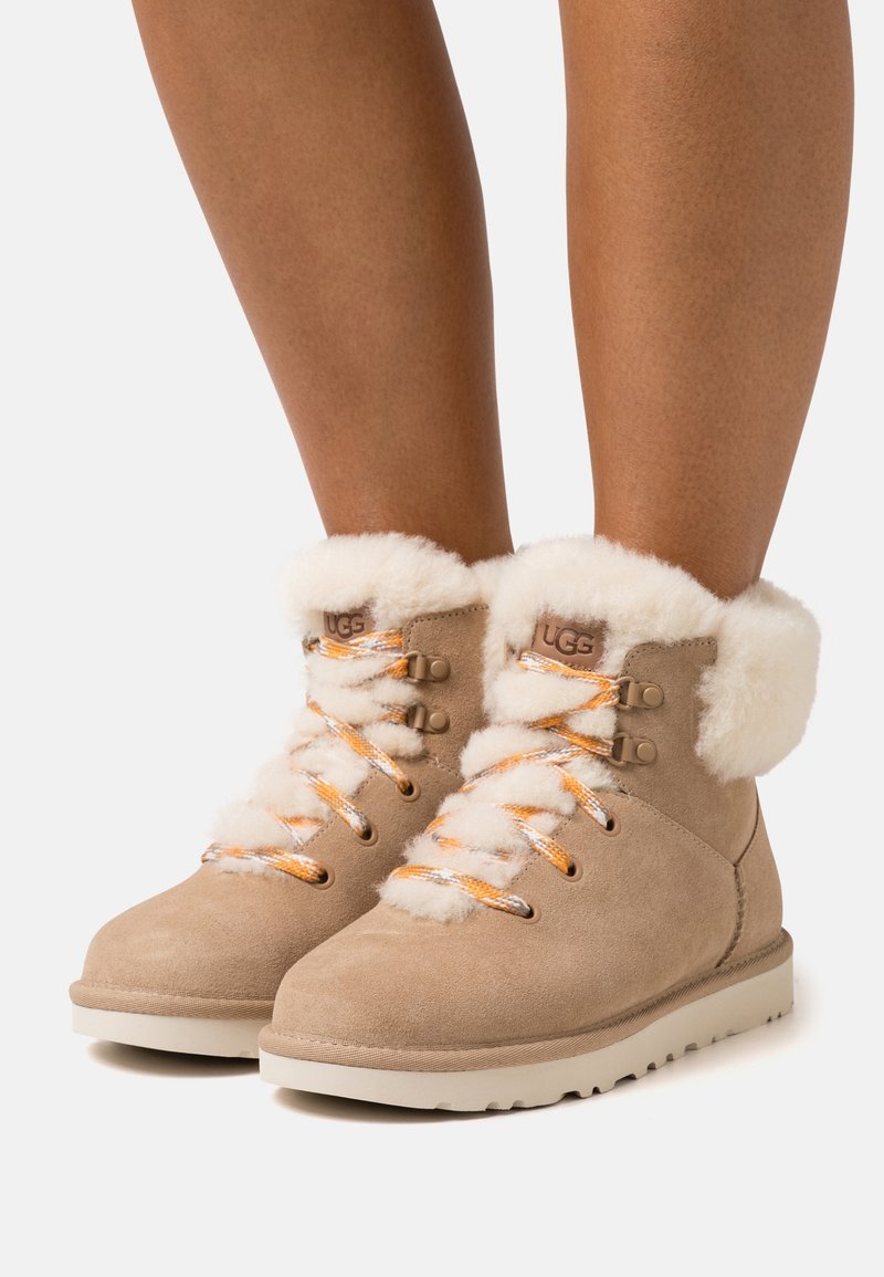 Braune Wildleder-Stiefeletten mit Lammfellfutter, orange-weiß gemusterte Schnürsenkel und Metallösen. Fester Gummisohle mit strukturiertem Profil.