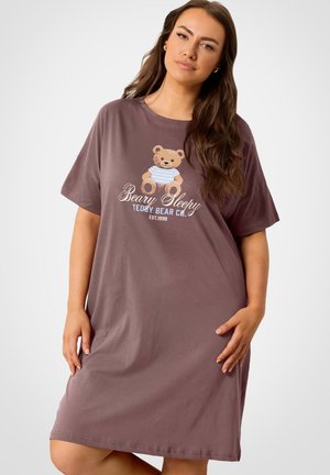 Nainen, joka pitää yllään löysää violettia yöpaitaa, jossa on nallekarhu-kuvio ja teksti "Beary Sleepy Teddy Bear Co. EST.1998", seisoo valkoisen taustan edessä.