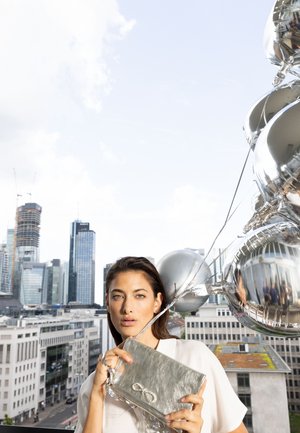 Silver metallic clutch bag with an infinity symbol, held in one hand. The background features urban architecture and balloon accents.