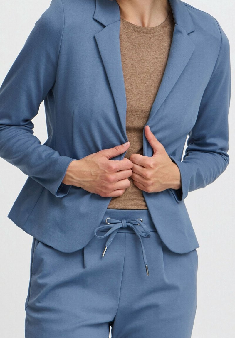 Personne ajustant un blazer bleu porté sur une chemise marron et un pantalon bleu assorti à cordon, sur un fond uni.