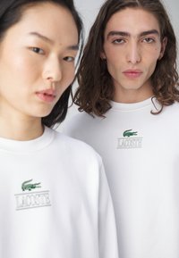 Fehér pamut pulóverek zöld hímzett Lacoste logóval és felirattal, kerek nyakkal és laza fazonnal, sima textúrával.