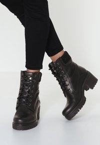 Bottes chevilles en cuir noir avec un talon épais, un design à lacets, des accents matelassés et une semelle en caoutchouc. Finition lisse et détails texturés.