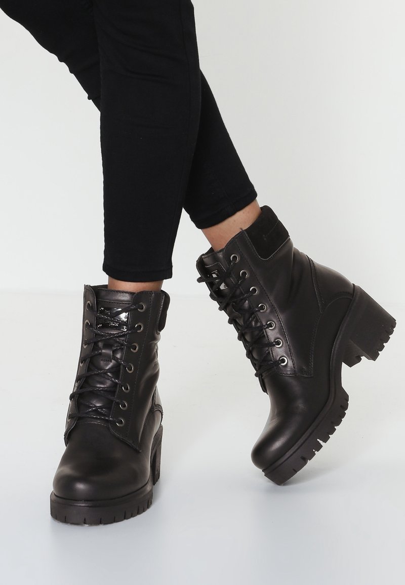Bottes chevilles en cuir noir avec un talon épais, un design à lacets, des accents matelassés et une semelle en caoutchouc. Finition lisse et détails texturés.