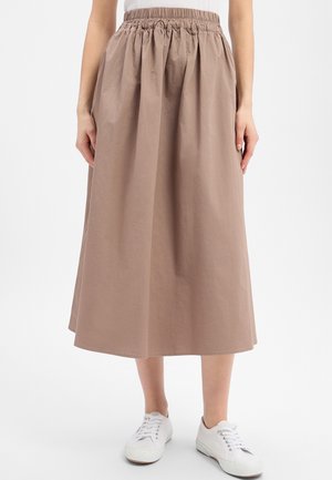 Kobieta ubrana w beżową, zebrą spódnicę midi z wysokim stanem, w zestawieniu z białymi trampkami, stojąca na jednolitym tle.