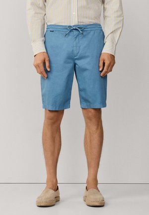 Hombre con pantalones cortos azul claro hasta la rodilla con cordón, camiseta de manga larga a rayas y zapatos beige sin cordones, de pie contra un fondo liso.